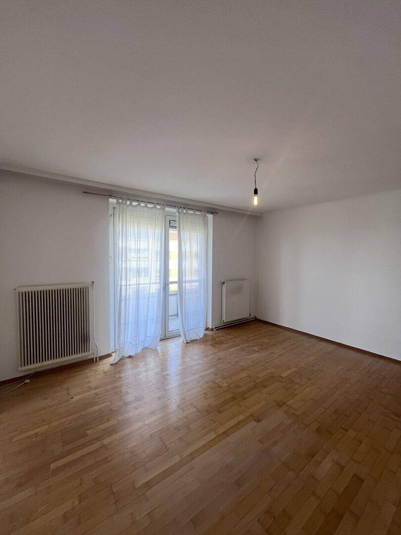 Geräumiges, leeres Wohnzimmer mit Parkettboden und Zugang zum Balkon.