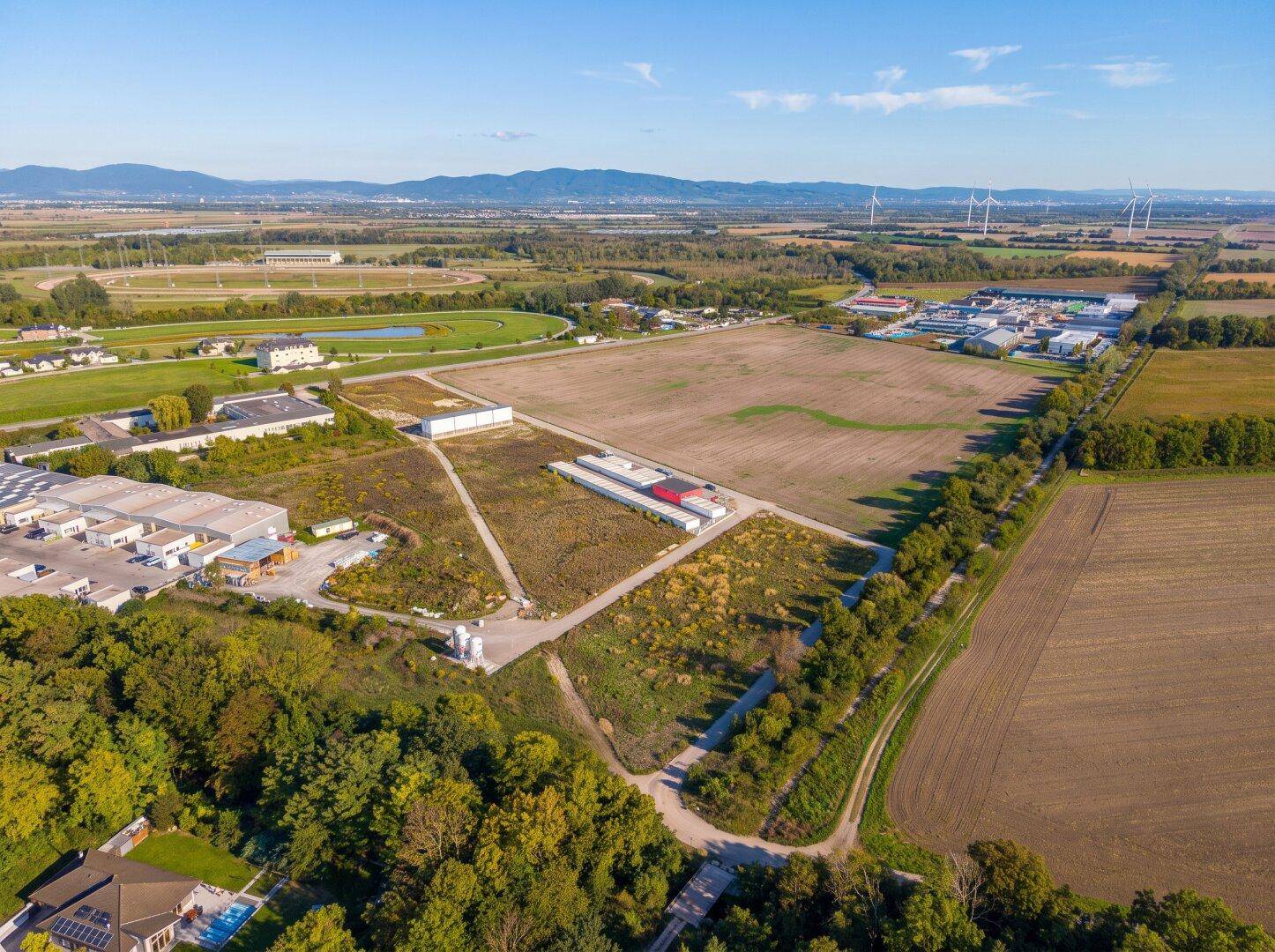 Weitläufige Luftaufnahme eines großen Grundstücks mit umliegenden Feldern, Industrie und Windrädern am Horizont.