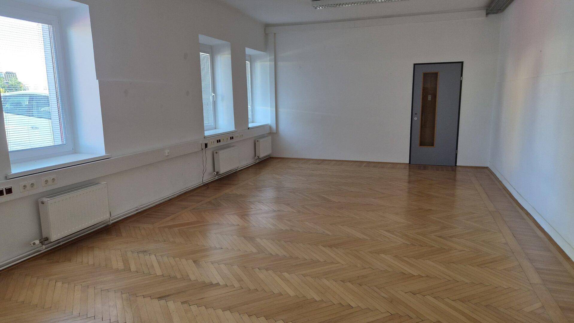 Heller, leerer Raum mit weiß gestrichenen Wänden, Fischgrätparkett und mehreren Fenstern.