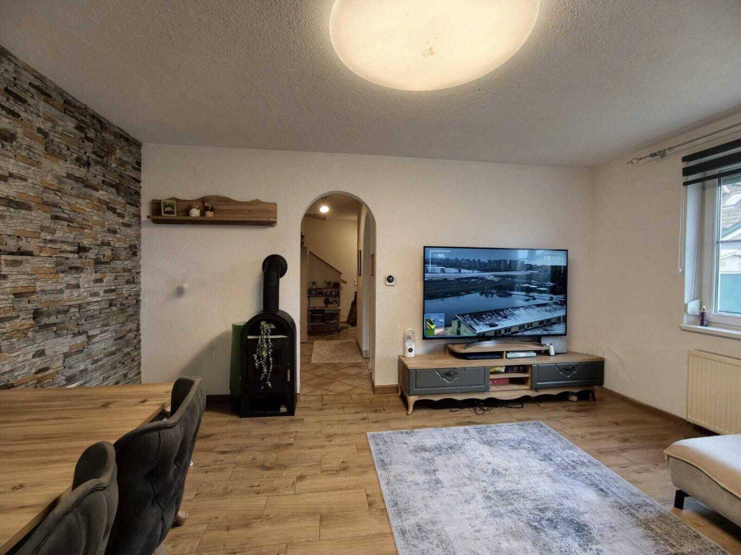 Wohnbereich mit Holzboden, Steinwand, Fernseher und Zugang zum Flur.