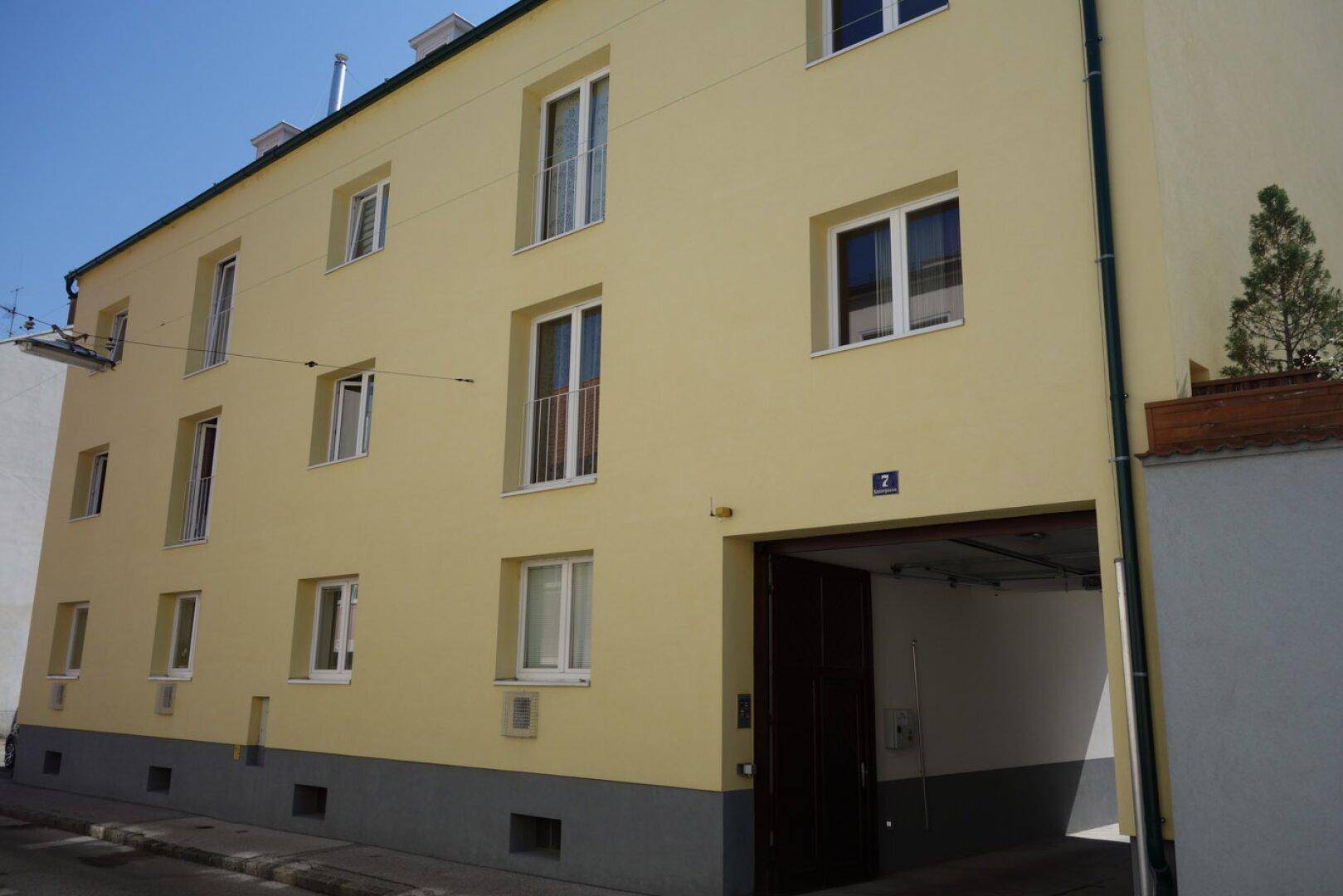 Gelbe Gebäudefassade mit mehreren Fenstern, französischen Balkonen und einer Garageneinfahrt.