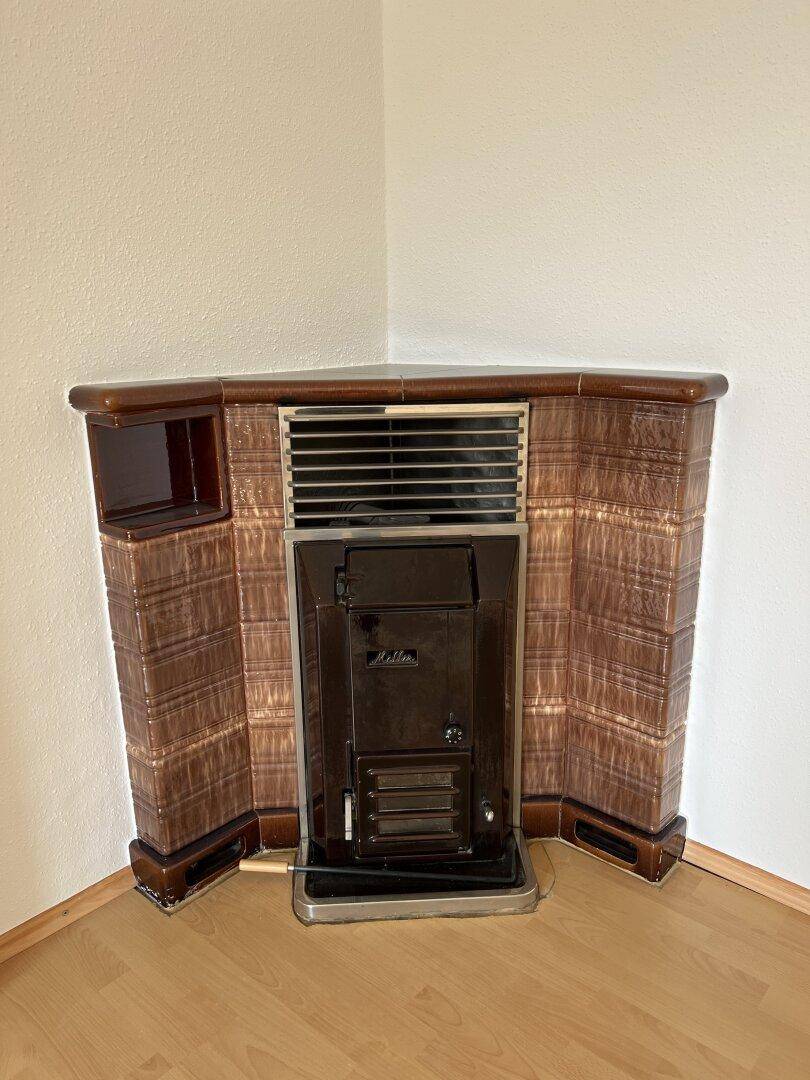Gemütlicher Kachelofen in der Ecke eines Raumes mit hellem Laminatboden und weißen Wänden.