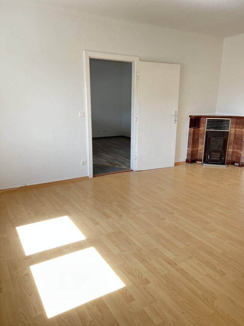 Heller Wohnraum mit Laminatboden, weißen Wänden und einem traditionellen Kachelofen, durchflutet von Tageslicht.