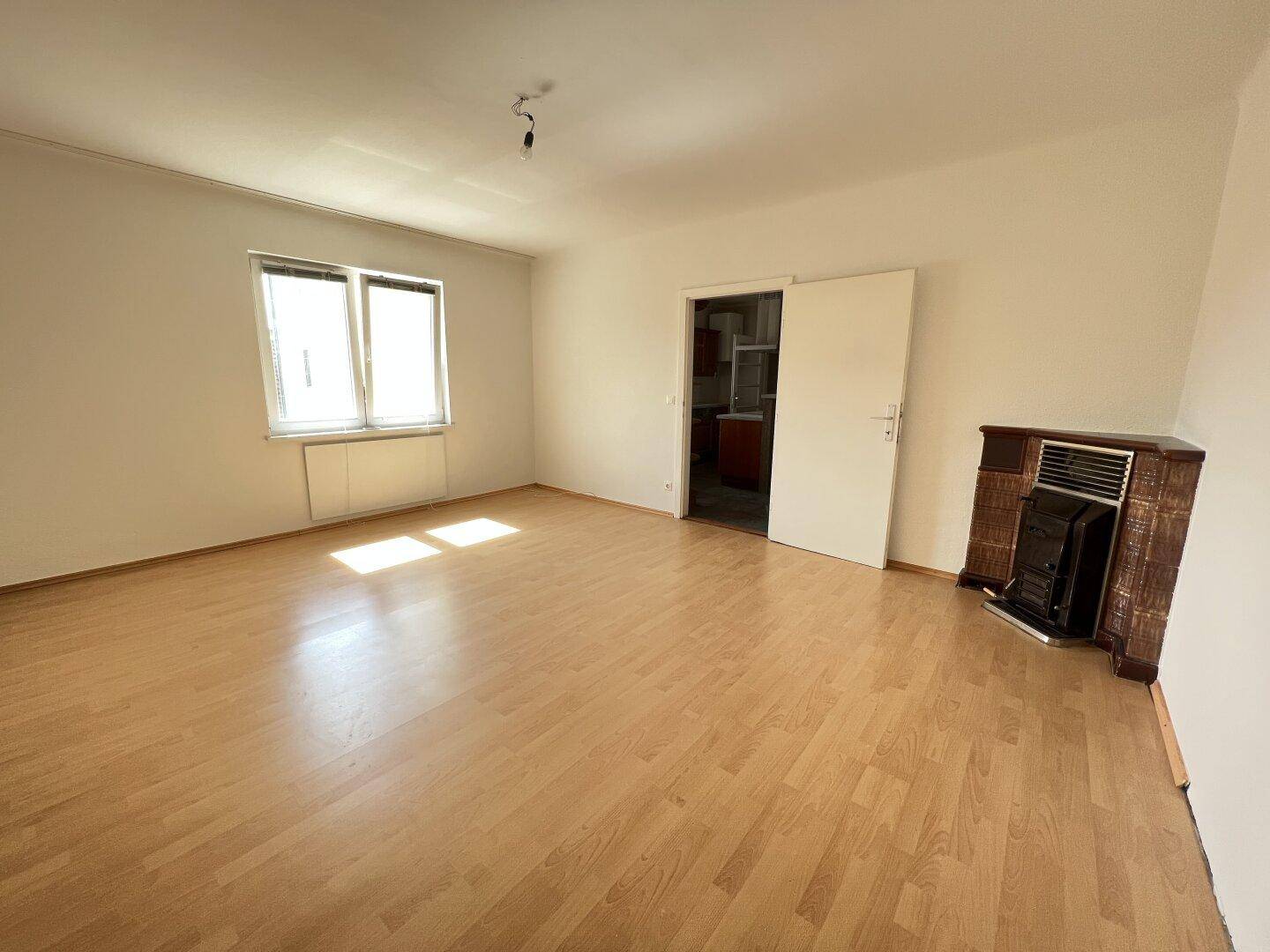 Geräumiges Wohnzimmer mit hellem Laminatboden, großen Fenstern und einem traditionellen Kachelofen.