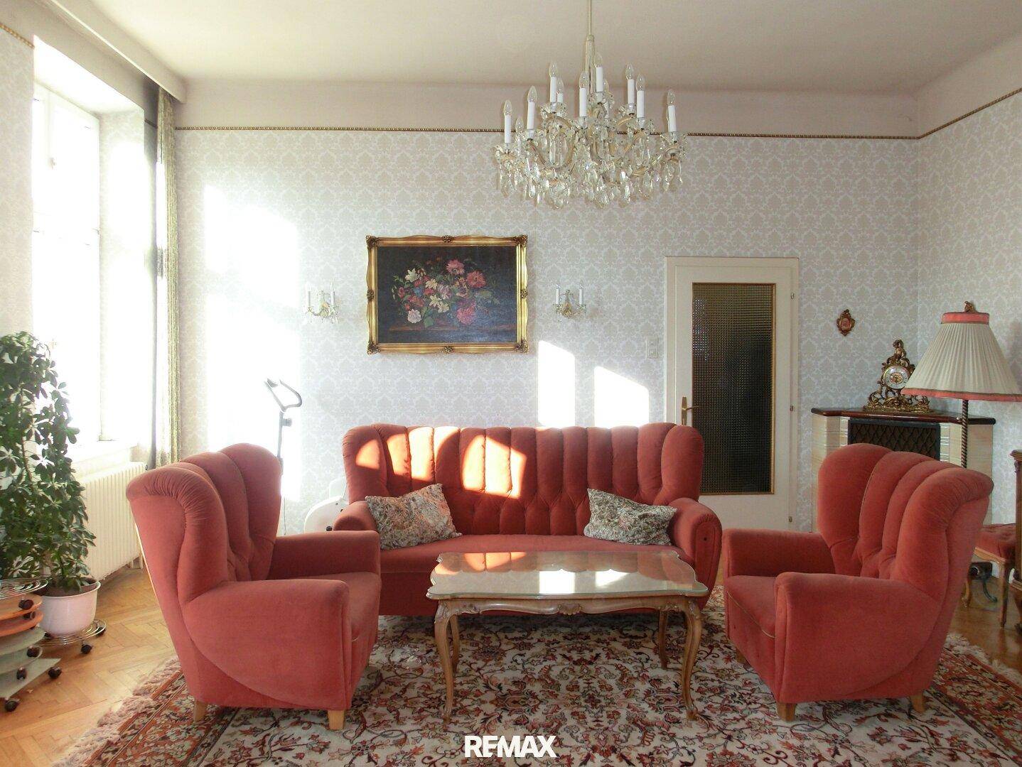 Gemütliches Wohnzimmer mit rotem Sofa, Sesseln und einem klassischen Kronleuchter.