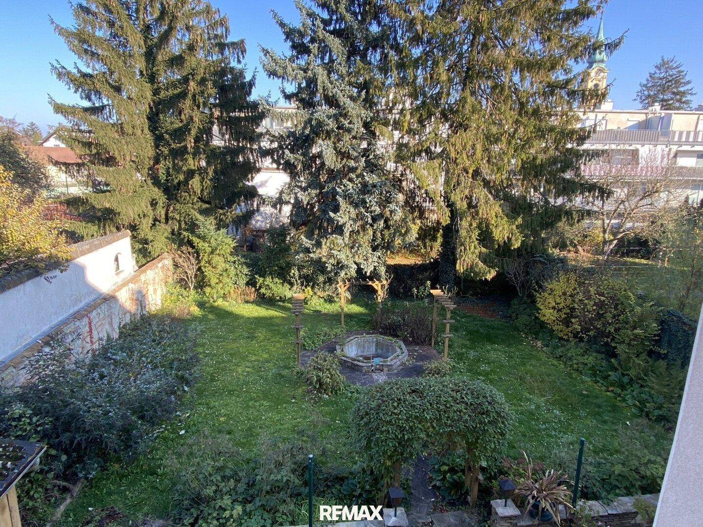 Großer Garten mit Rasenfläche, hohen Bäumen und einem dekorativen Brunnen.
