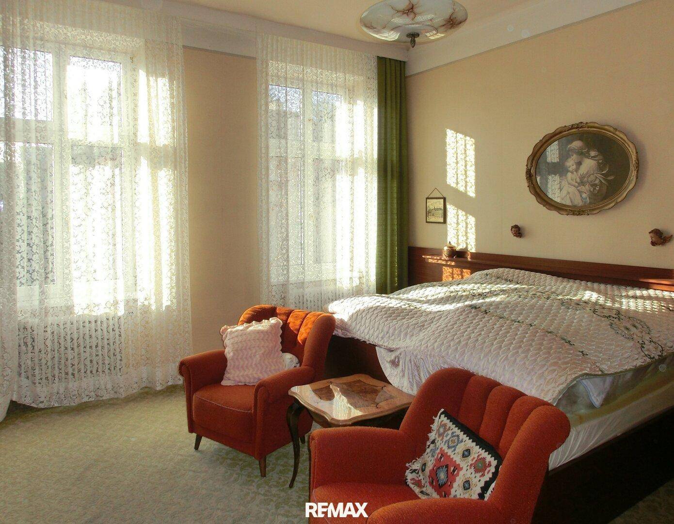 Schlafzimmer mit Doppelbett, zwei Sesseln und viel Tageslicht durch große Fenster.