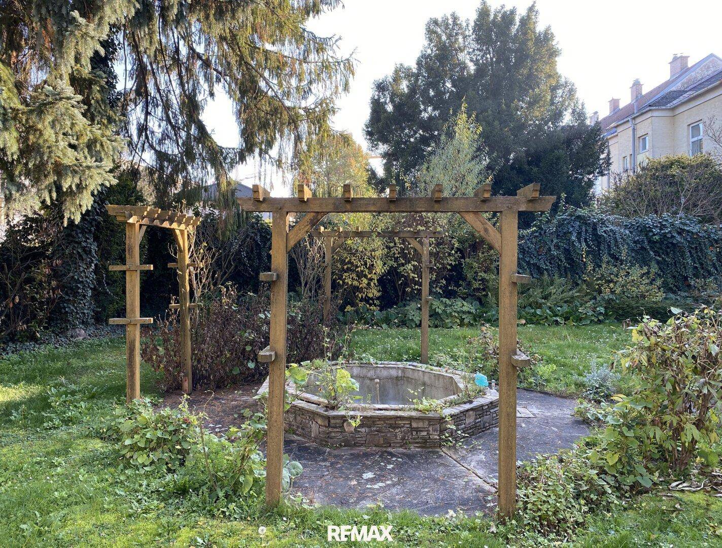 Gartenbereich mit Pergola, Brunnen und üppiger Vegetation.