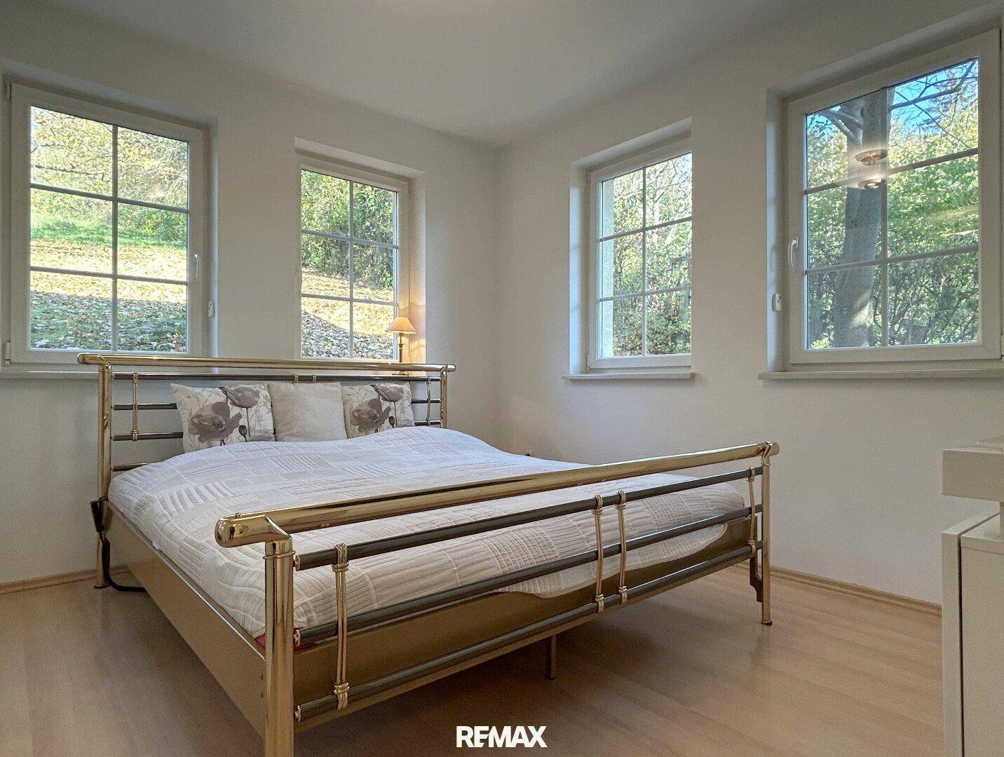 Helles Schlafzimmer mit Metallbett, Holzfußboden und Fenstern mit Blick ins Grüne.