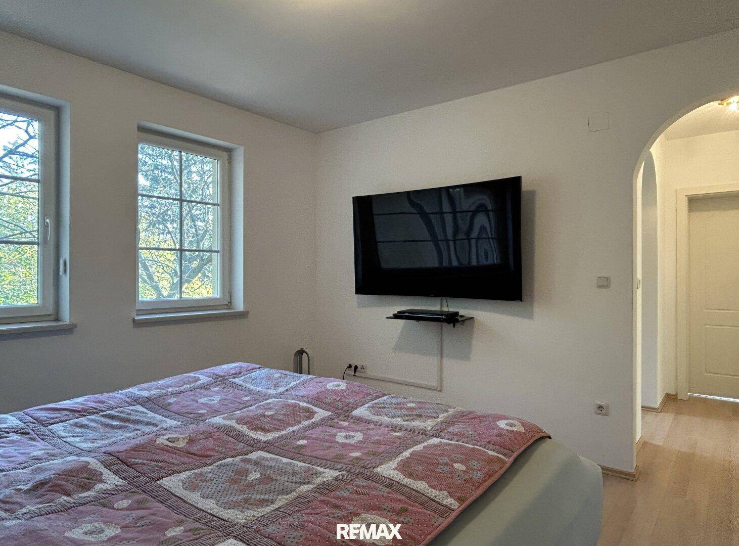 Schlafzimmer mit Wandfernseher und Zugang zu einem weiteren Raum durch einen Bogen.