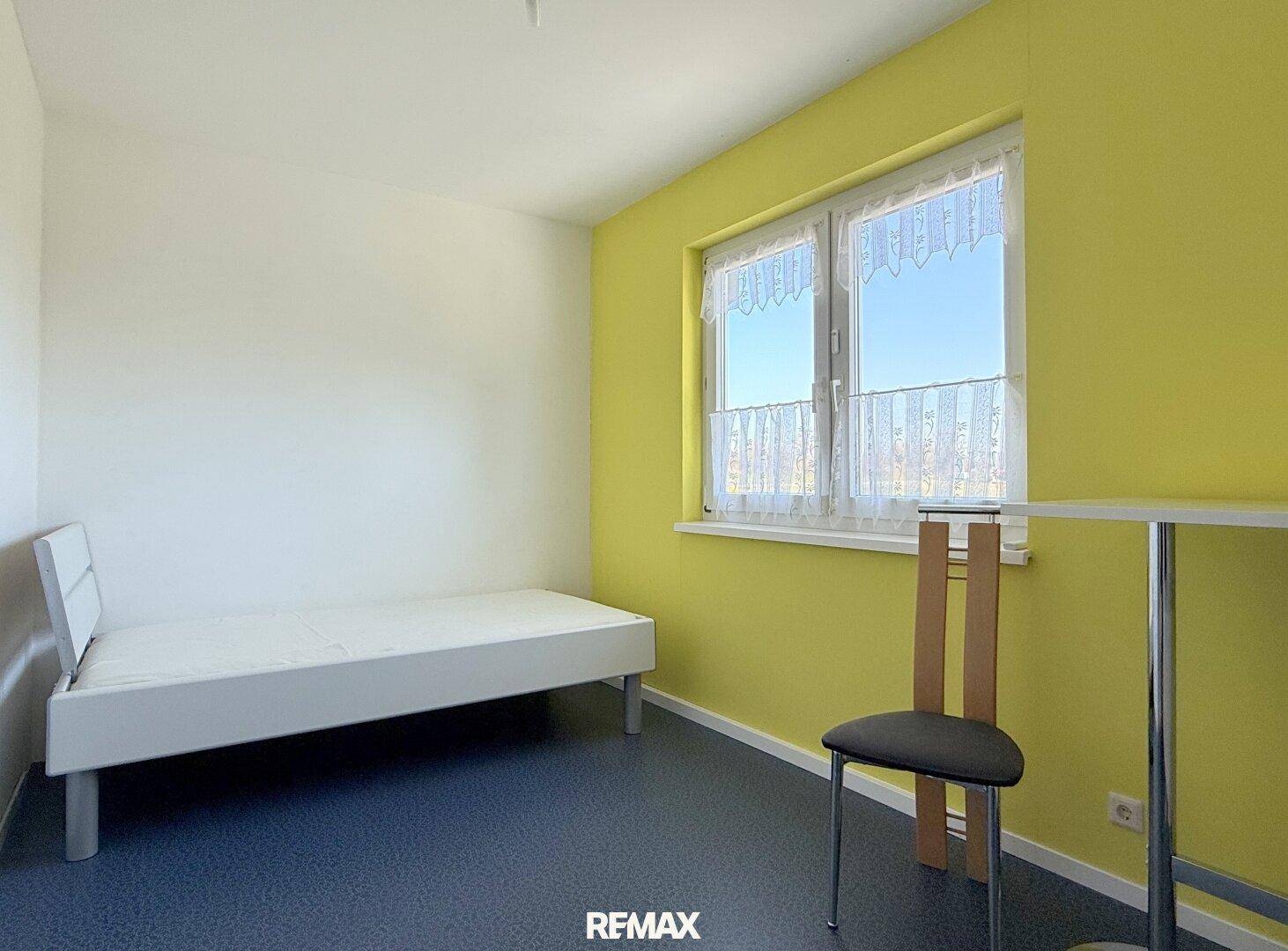 Helles Zimmer mit Einzelbett, gelber Wand und Fenster mit Blick ins Freie.