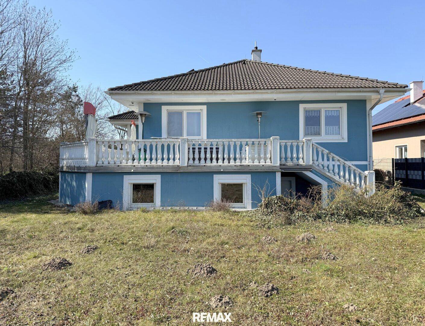 Freistehendes blaues Haus mit weißer Terrasse und Balustrade, umgeben von einem weitläufigen Garten.