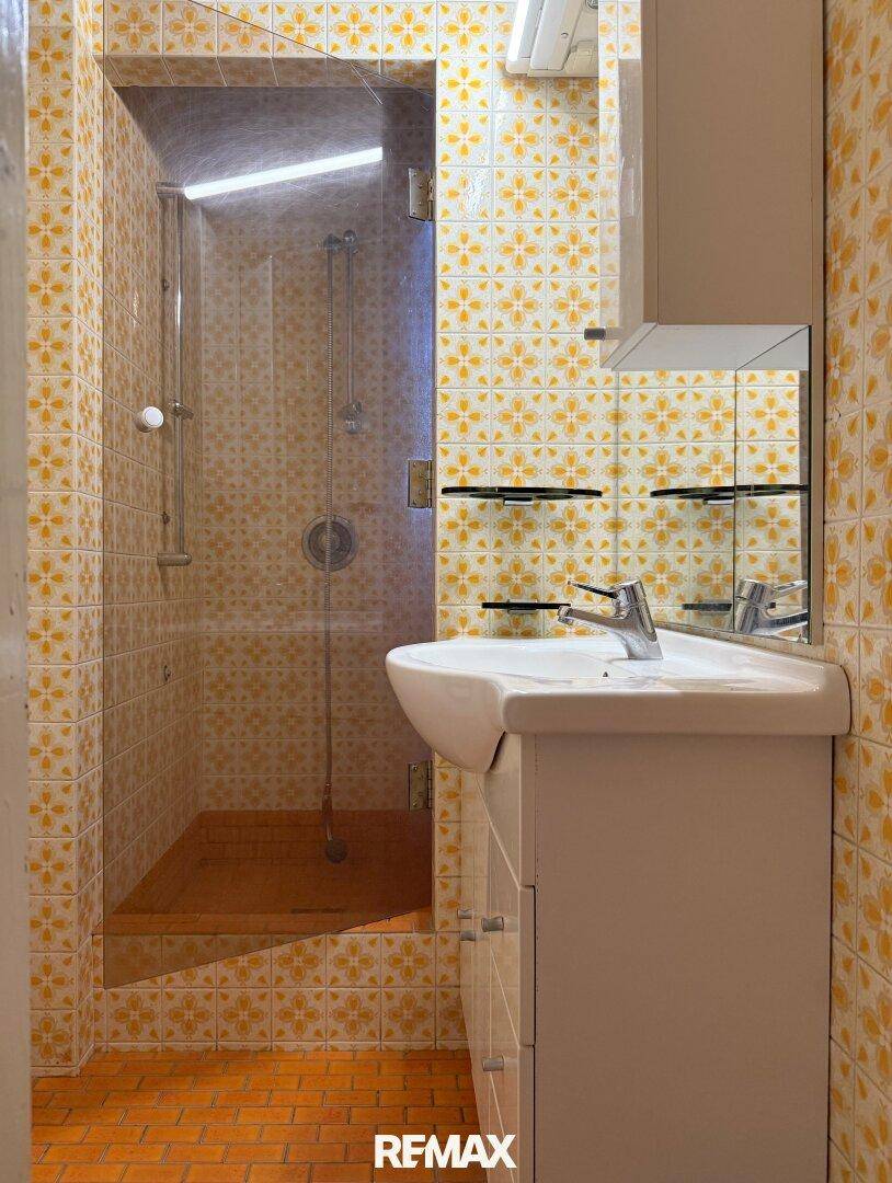 Badezimmer mit gelb-weiß gemusterten Fliesen, Einzelwaschbecken und einer Duschkabine.