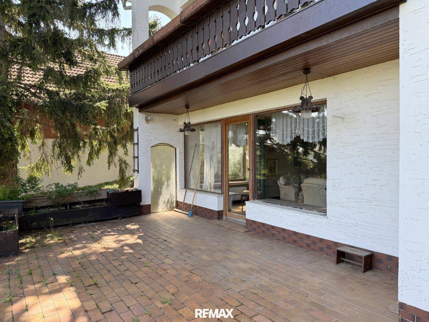 Gepflasterte Terrasse vor dem Haus mit großen Fenstern und einem darüberliegenden Balkon, umgeben von Bäumen.