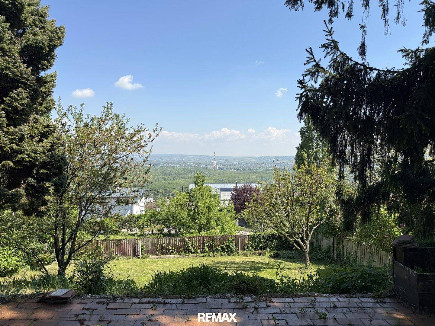 Weitläufiger Ausblick über einen grünen Garten und Bäume auf eine ferne Landschaft mit Gebäuden unter blauem Himmel.