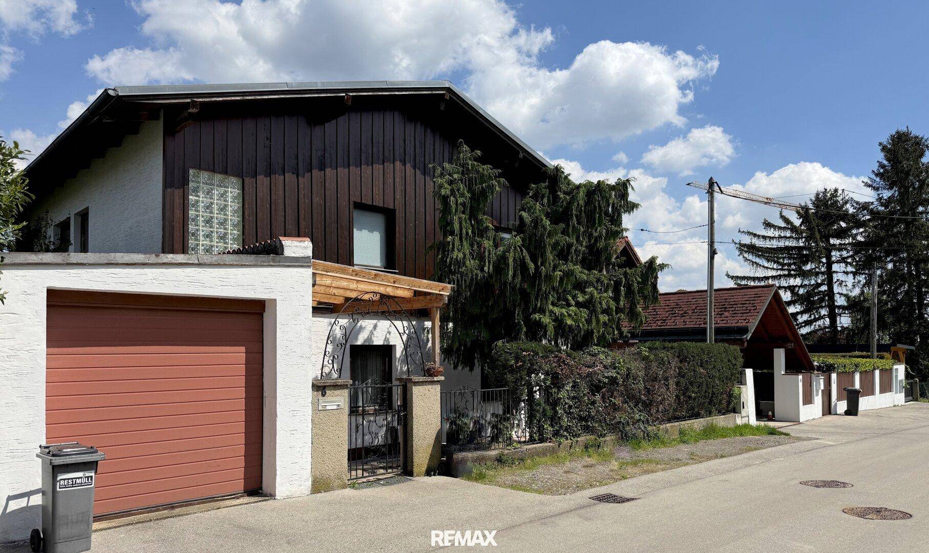 Außenansicht eines Hauses von der Straße mit Garage, weißer Fassade, Holzverkleidung und einem kleinen überdachten Eingangsbereich.