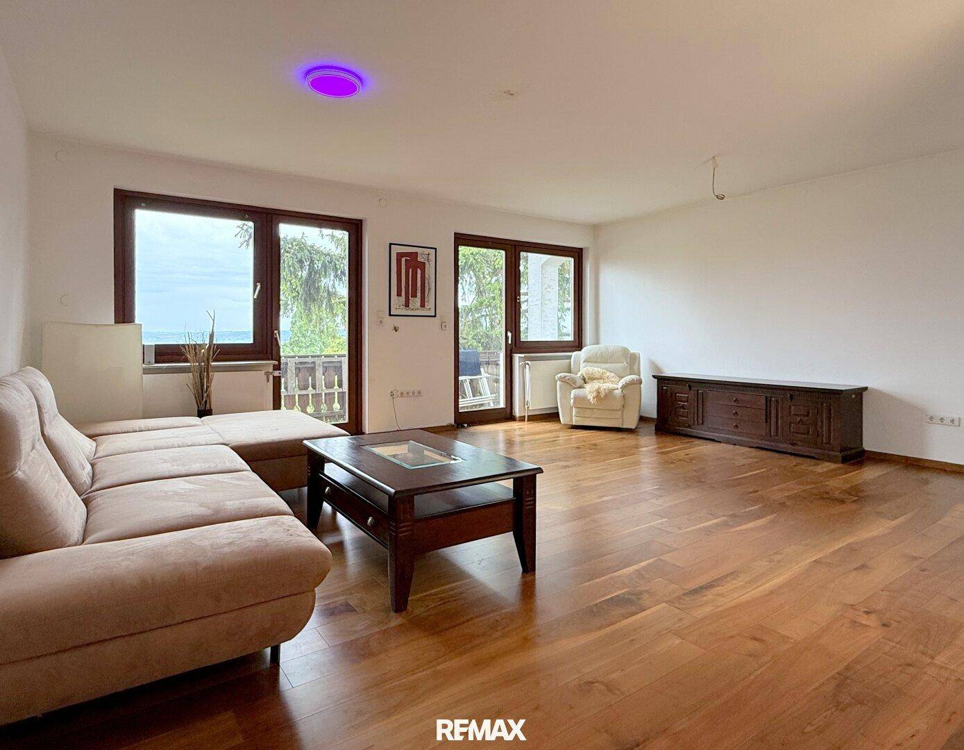 Helles Wohnzimmer mit Parkettboden, beiger Couch und großen Fenstern mit Zugang zum Balkon.