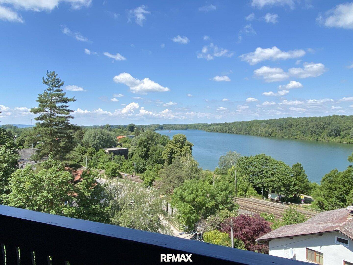 Weitläufiger Blick von einem Balkon auf den See und die grüne Landschaft unter blauem Himmel.