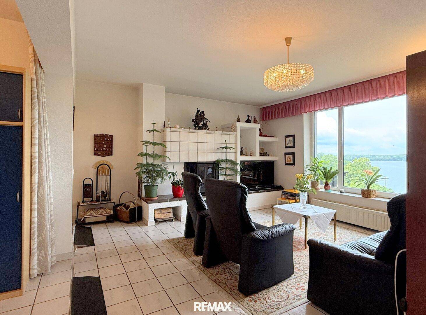 Geräumiges Wohnzimmer mit Kamin, Fliesenboden und großen Fenstern mit Seeblick.