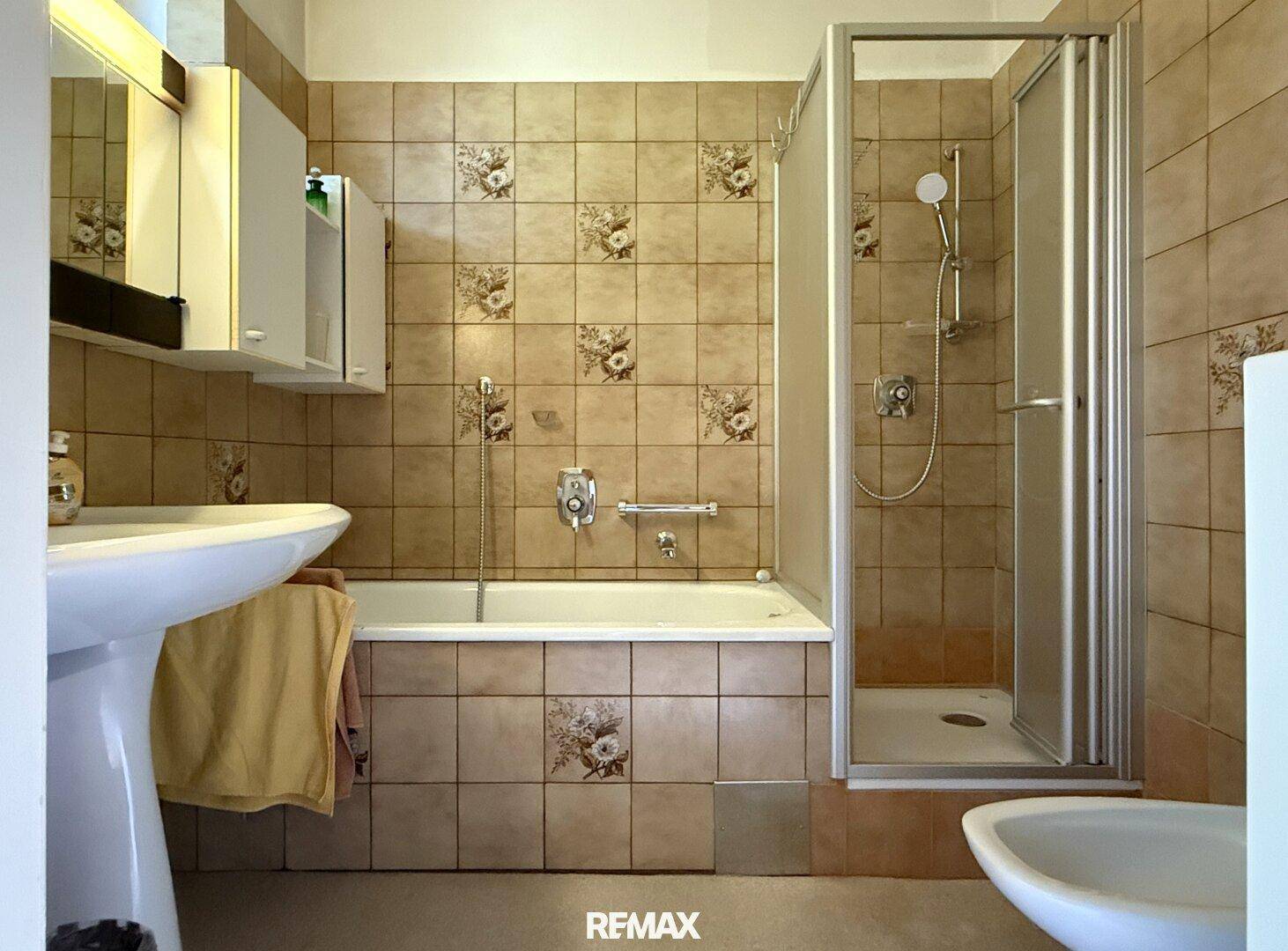 Badezimmer mit Badewanne, Dusche und floral gemusterten Fliesen im 80er-Jahre-Stil.