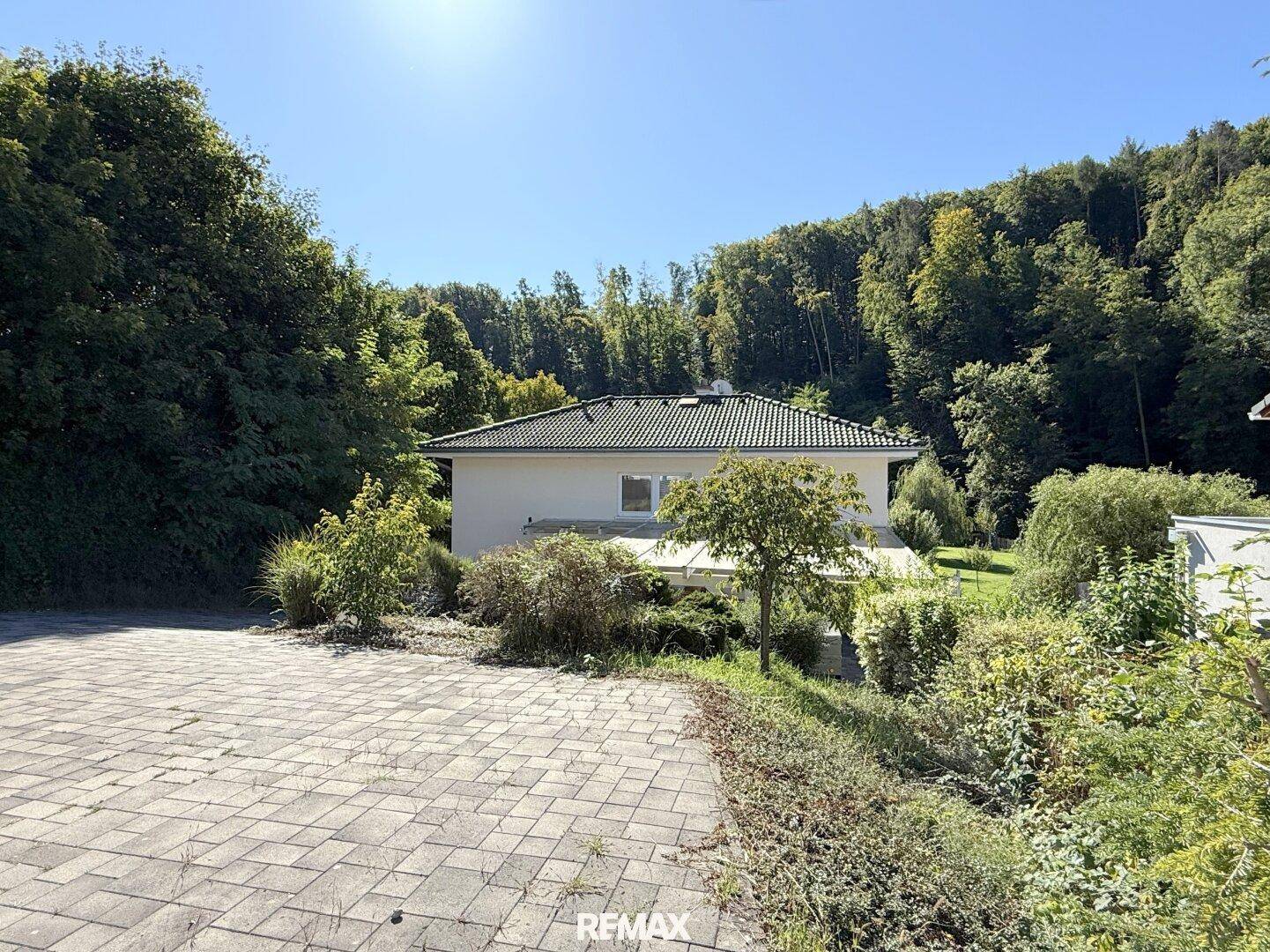 Einfamilienhaus mit gepflasterter Auffahrt, umgeben von üppigem Grün und Wald in sonniger Lage.