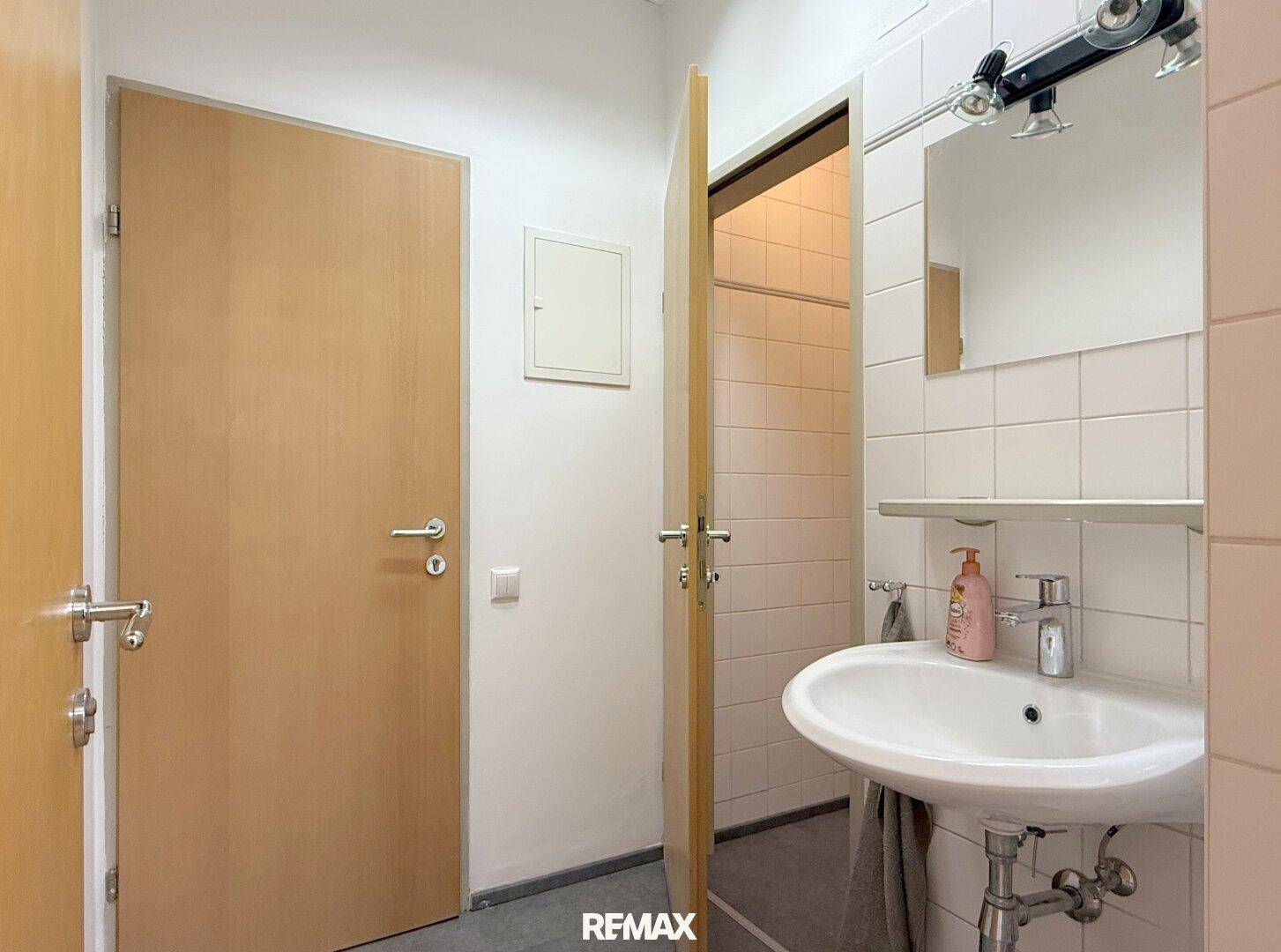 Funktionales Badezimmer mit weißem Waschbecken, Spiegel und hellen Fliesen an den Wänden.