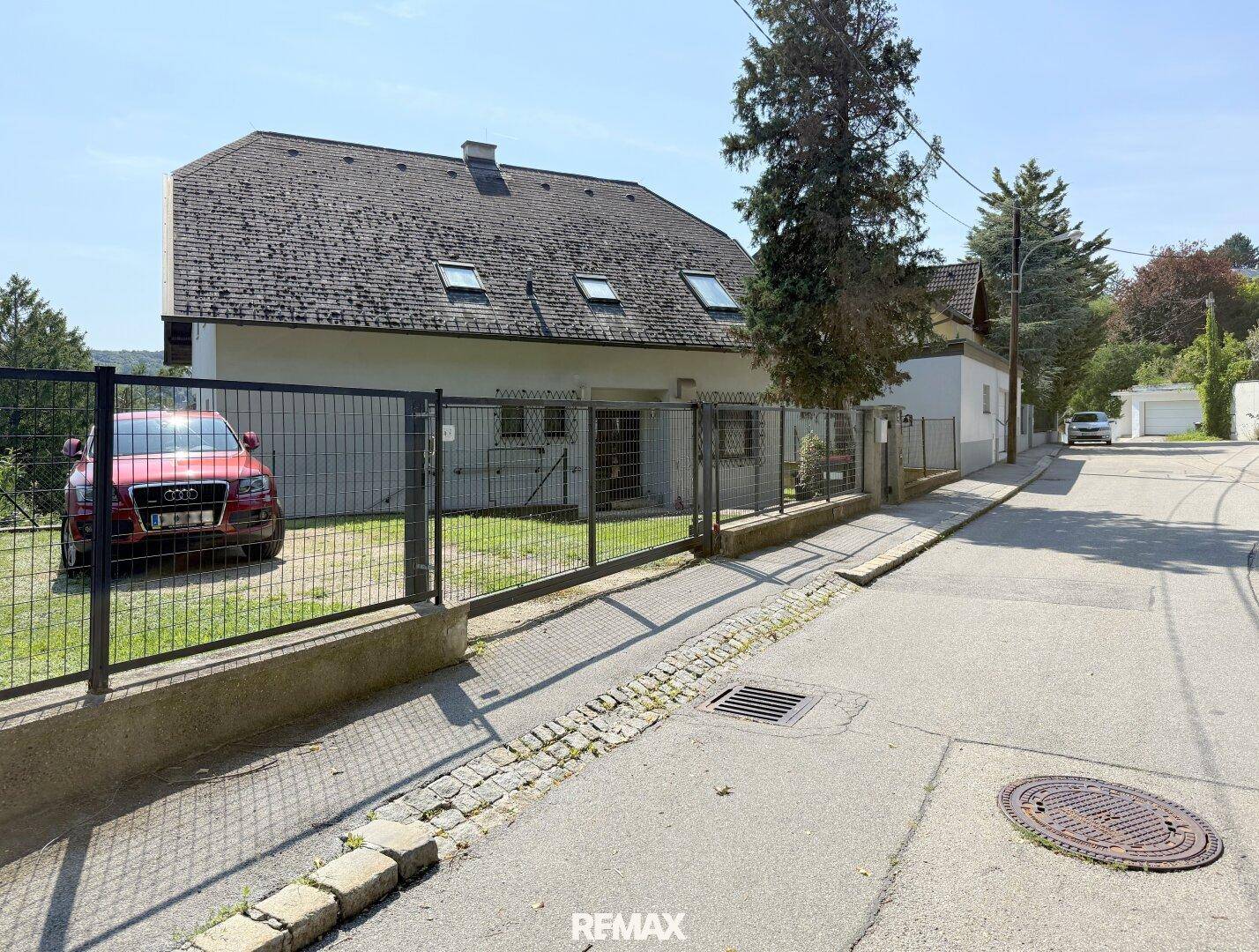 Ansicht des Hauses von der Straße mit Einfahrt und Zaun, umgeben von Grünflächen.