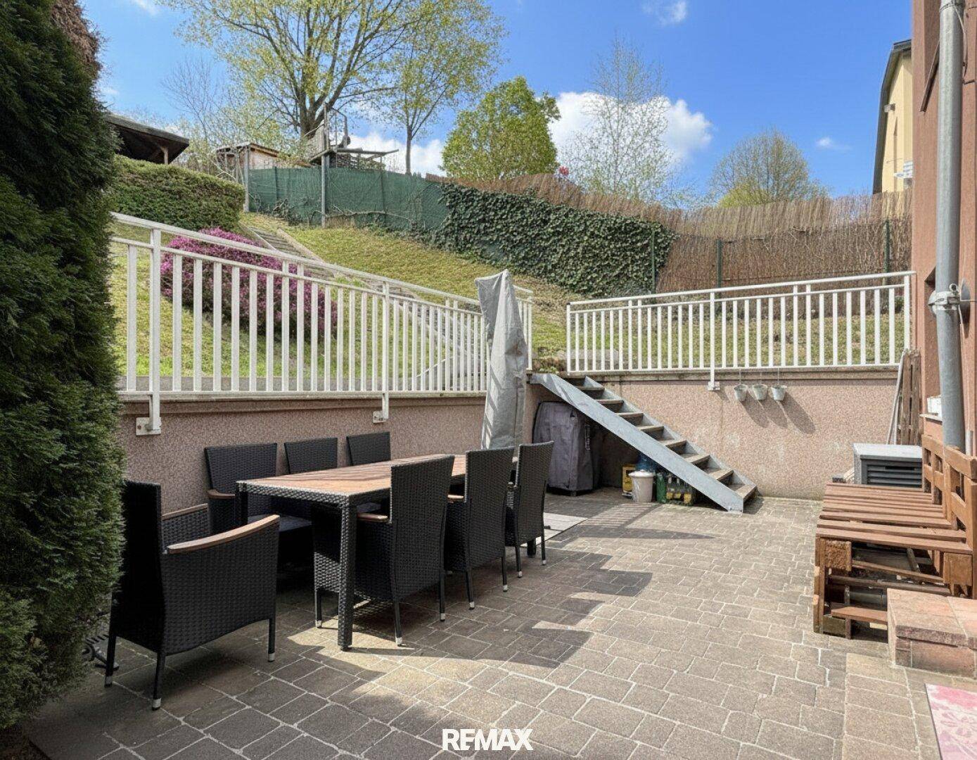 Gepflasterte Terrasse mit Esstisch und Stühlen, umgeben von einer grünen Böschung.