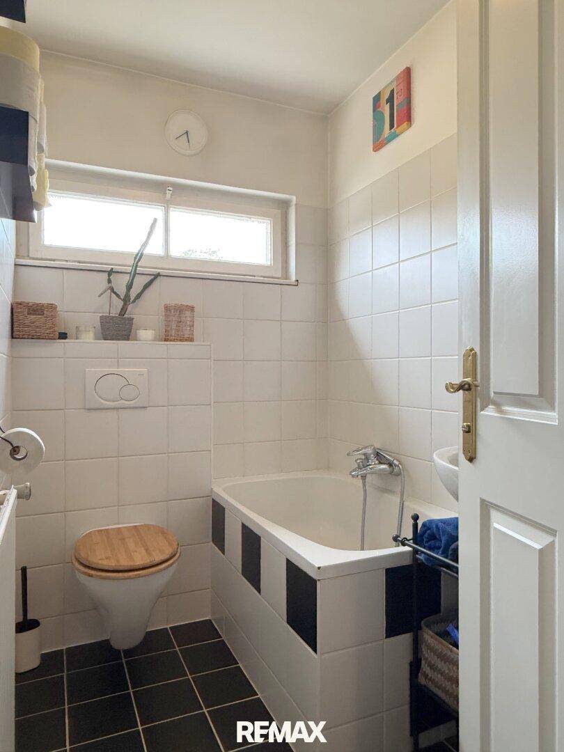 Kleines Badezimmer mit Badewanne, WC und Fenster, gefliest in Weiß und Schwarz.