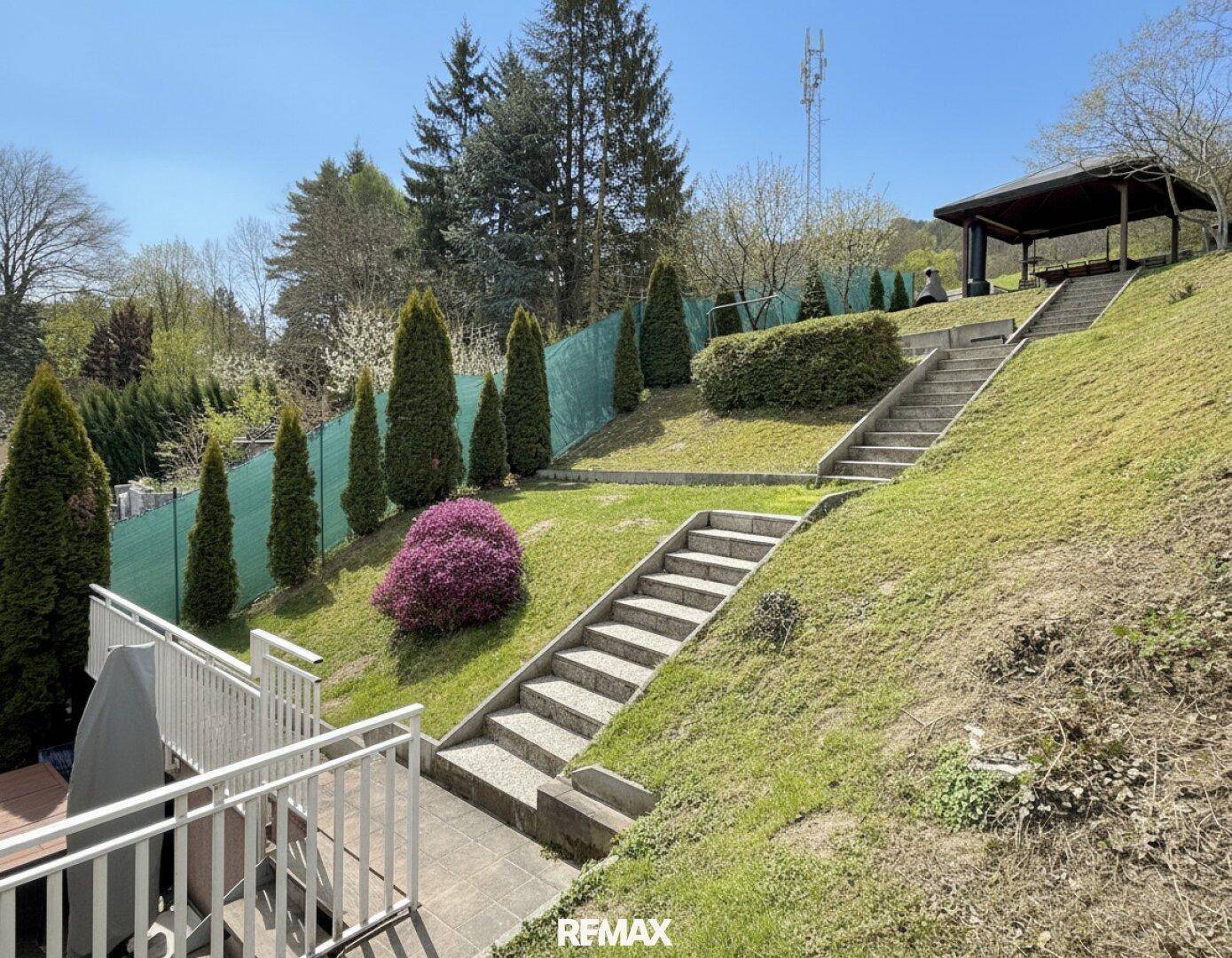 Weitläufiger, gestufter Garten mit mehreren Ebenen, Steintreppen und einem Pavillon unter blauem Himmel.