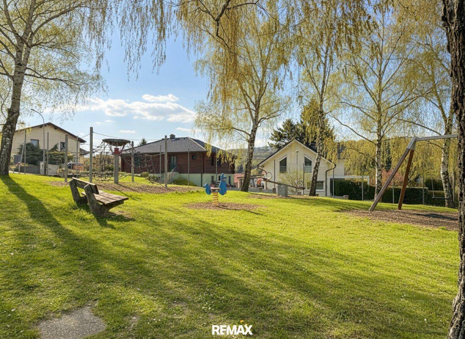 Sonniger Spielplatz mit Schaukeln, Rutsche und einer Holzbank, umgeben von Bäumen und Häusern.