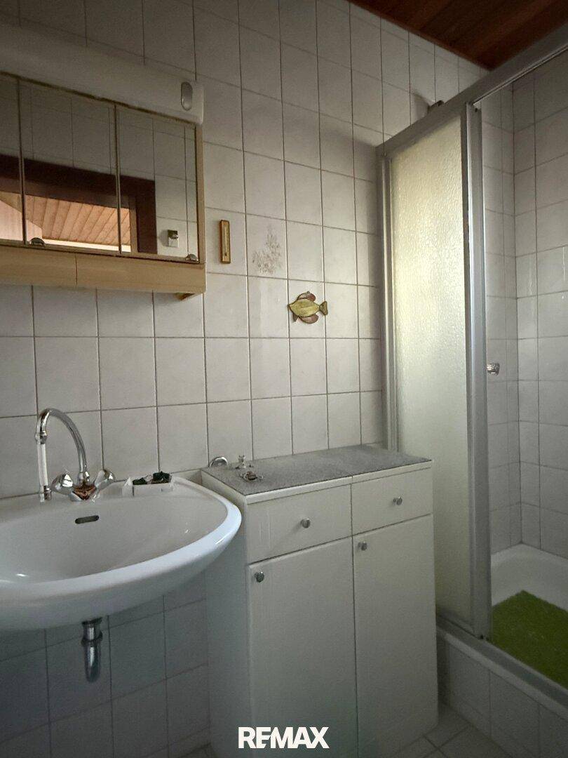 Badezimmer mit weißen Fliesen, Waschbecken und einer Duschkabine, im Stil der 80er Jahre.