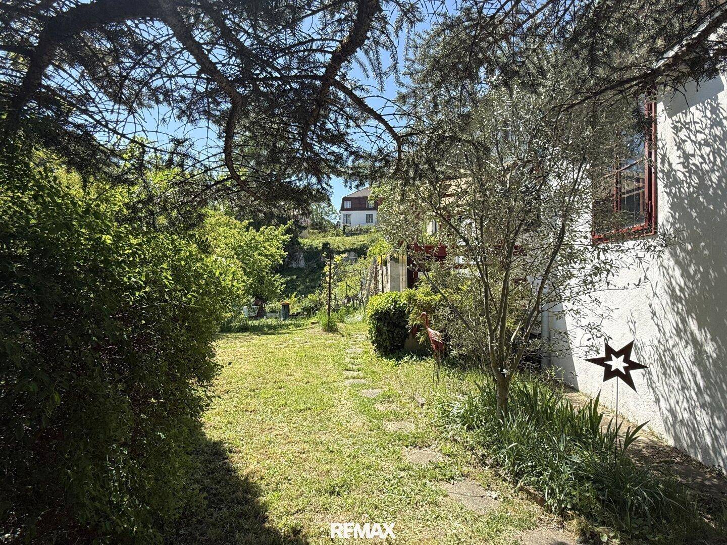 Gartenweg entlang des Hauses, umgeben von üppigem Grün und Bäumen unter strahlendem Himmel.