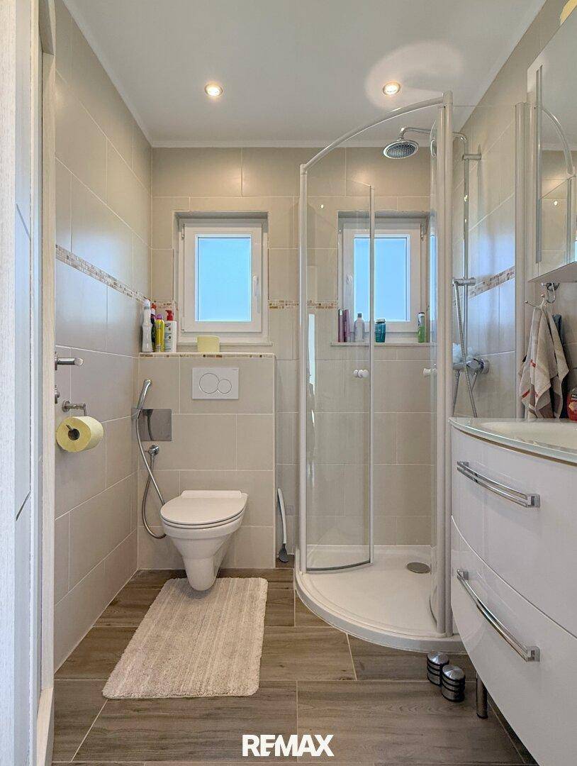 Modernes Badezimmer mit Dusche, WC, Waschbecken und zwei Fenstern für Tageslicht.