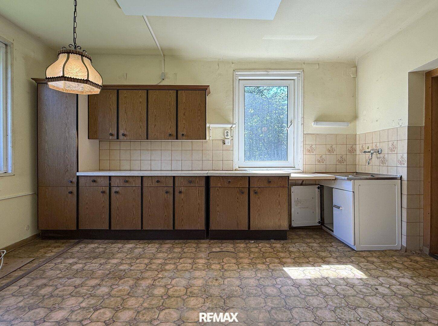 Ältere Küche mit Holzschränken, Spüle und einem Fenster, das viel Tageslicht hereinlässt.