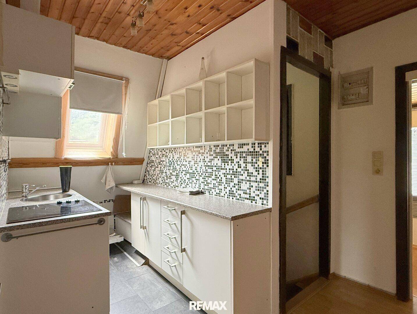 Kleine Küchenzeile mit Spüle, Kochfeld und Regalen unter einer Holzdecke.