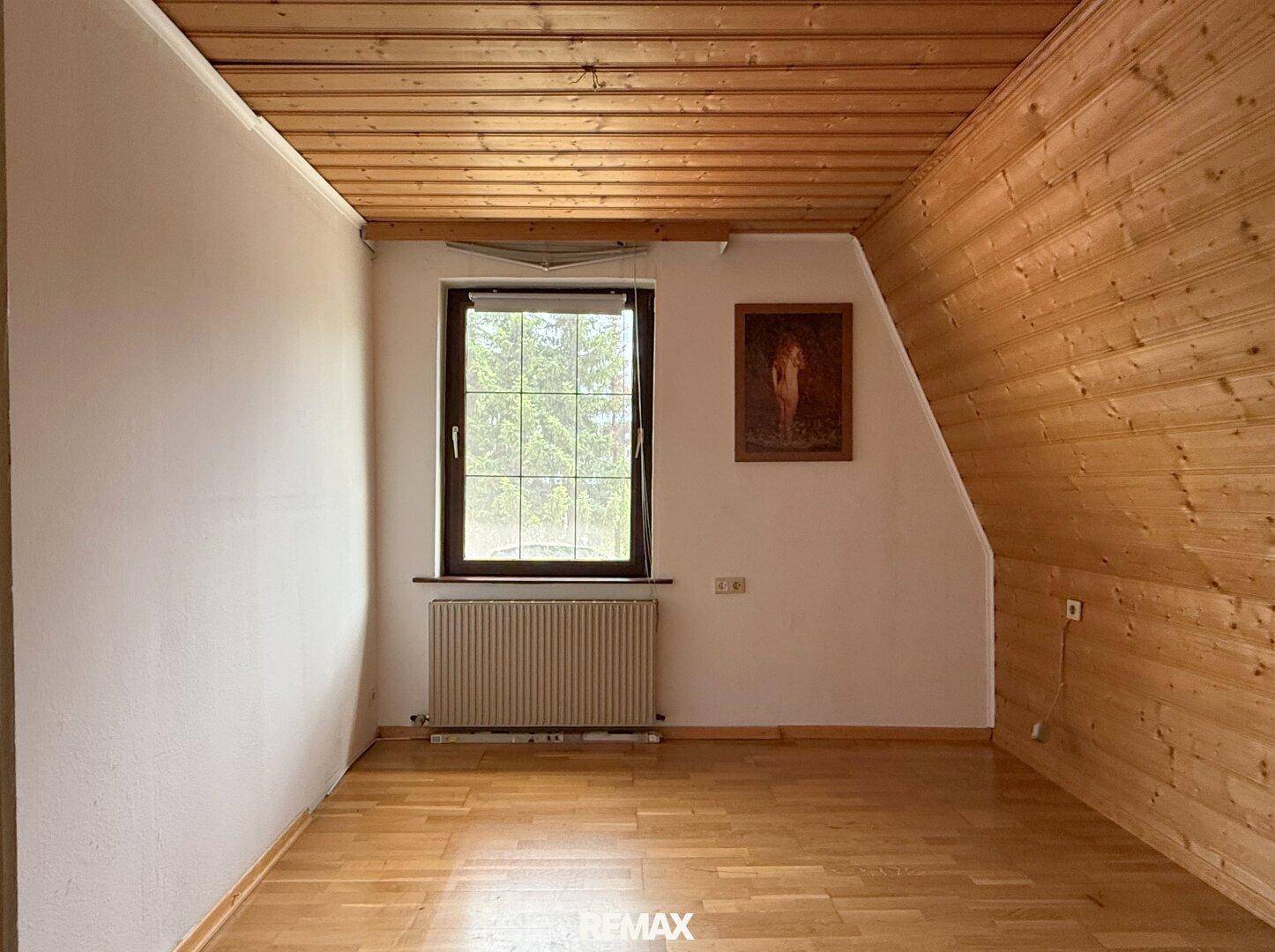 Dachzimmer mit Holzverkleidung, Parkettboden und Fenster mit Blick ins Grüne.