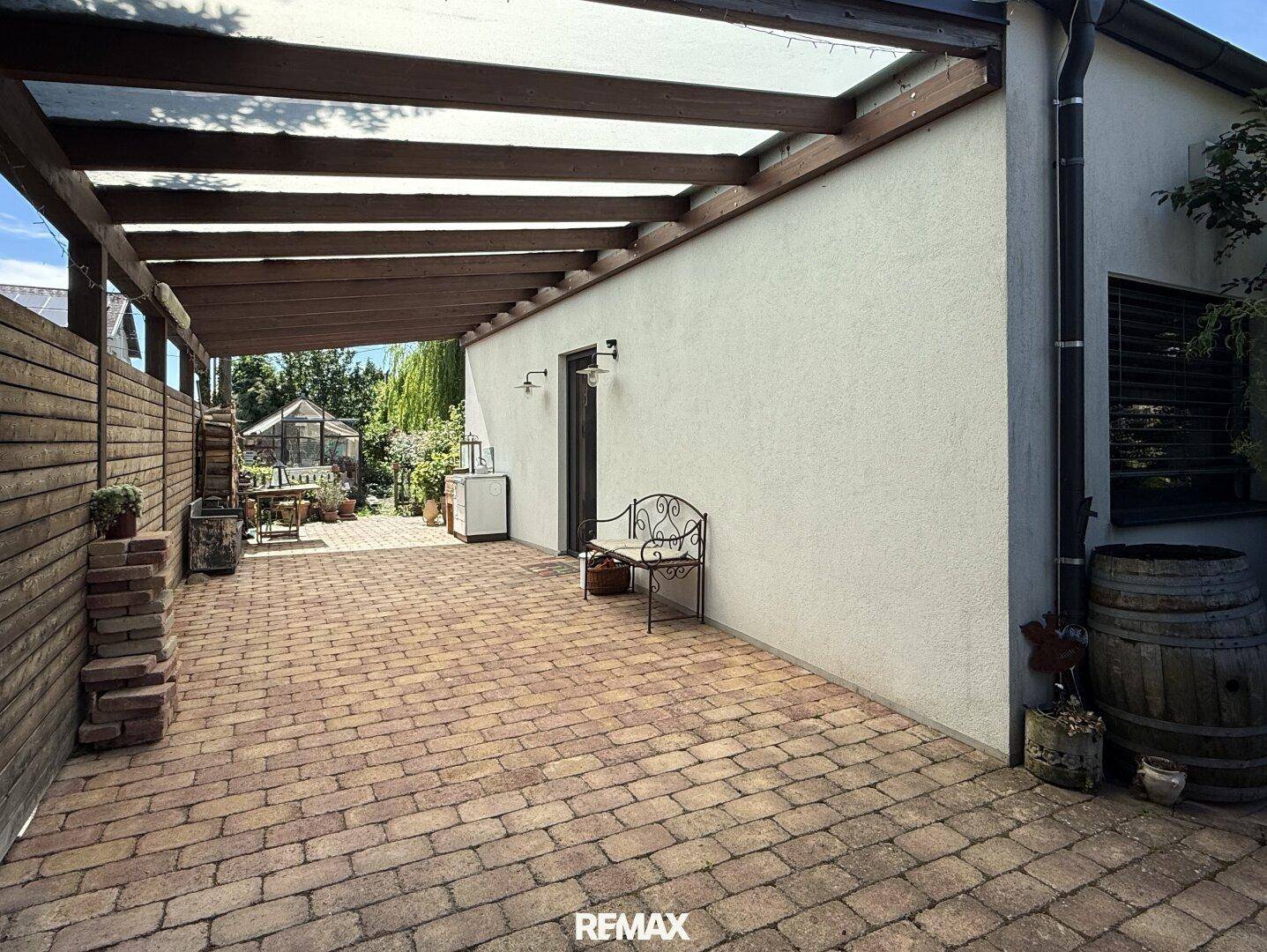 Geräumige überdachte Terrasse mit Zugang zum Garten und Blick auf ein Gewächshaus.