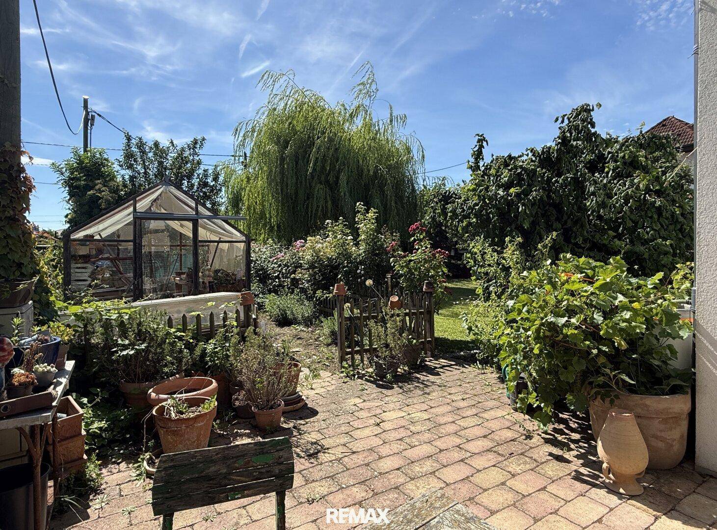 Gepflegter Garten mit Gewächshaus, gepflastertem Weg und üppiger Bepflanzung.