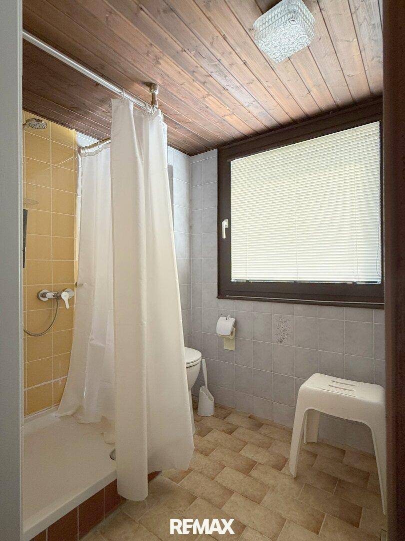 Einfaches Badezimmer mit Duschkabine, WC, Fenster und Holzdecke.