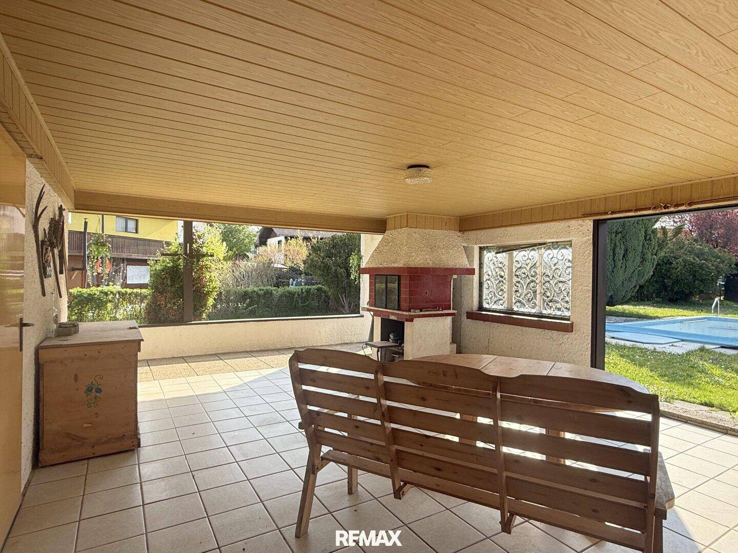 Überdachte Terrasse mit Fliesenboden, Holzbänken und einem gemauerten Grill.