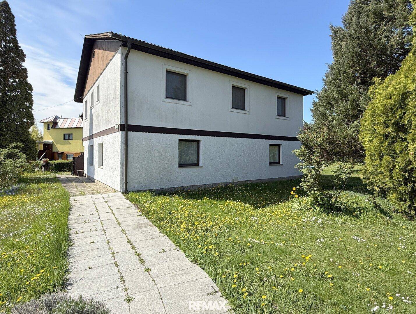 Seitenansicht des Hauses mit weißer Fassade, Gartenweg und blühender Wiese.