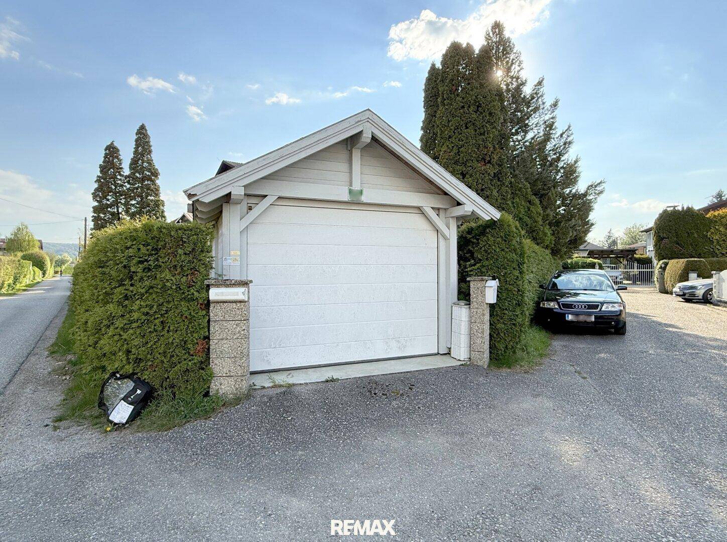 Garage mit weißem Tor, Zufahrt und einer dichten Hecke entlang der Straße.