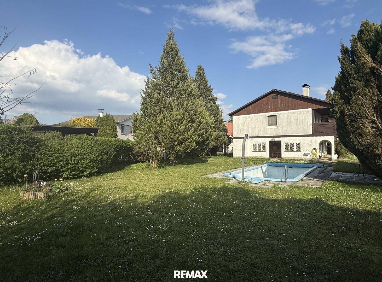 Großer Garten mit Rasenfläche, hohen Bäumen und einem Swimmingpool unter blauem Himmel.