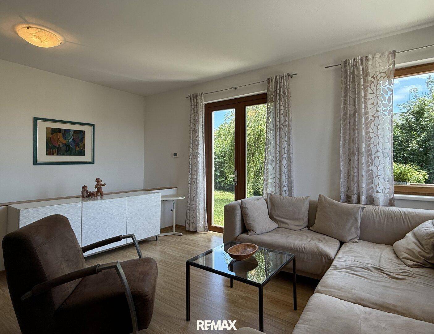Gemütliches Wohnzimmer mit beigem Sofa, Holzboden und großen Fenstern mit Blick in den Garten.