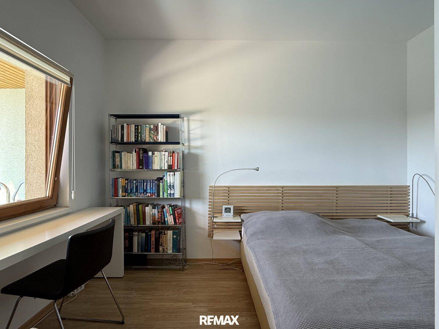 Schlafzimmer mit Holzbett, Schreibtisch und Fenster, das viel Tageslicht hereinlässt.