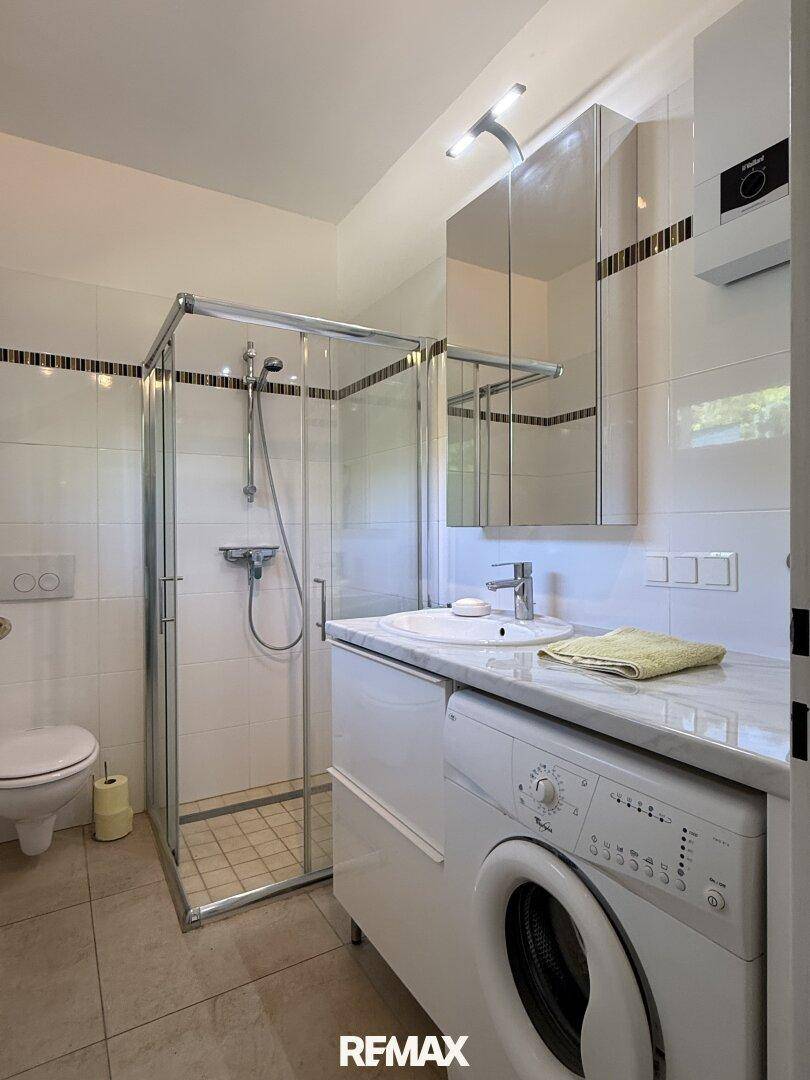 Modernes Badezimmer mit Dusche, Waschbecken und Waschmaschine, hell gefliest.