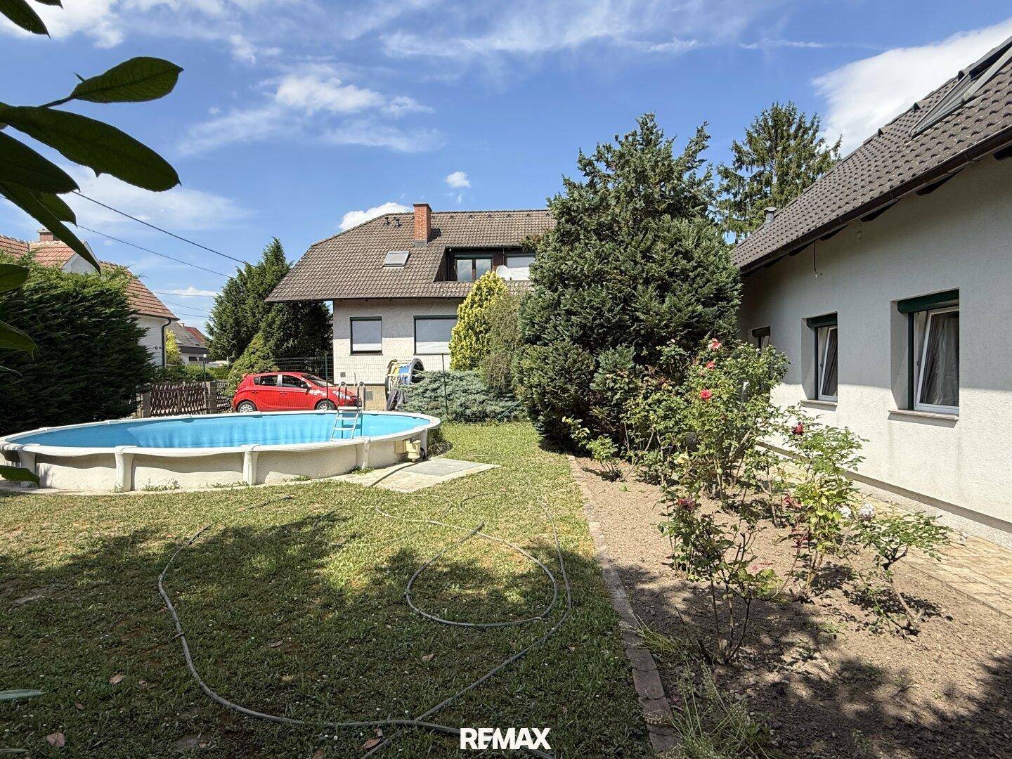 Weitläufiger Garten mit Aufstellpool und Rasenfläche, umgeben von Sträuchern und einem Wohnhaus im Hintergrund.
