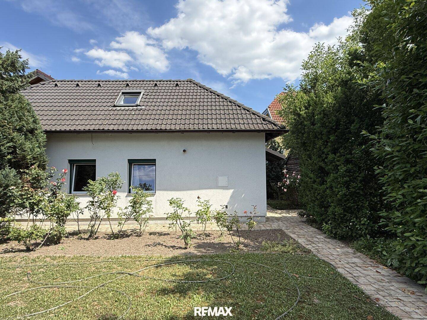 Seitenansicht eines Wohnhauses mit Ziegeldach, Dachfenster und gepflegten Rosenbeeten im Vorgarten.