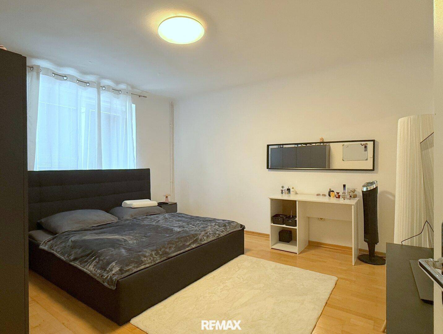 Schlafzimmer mit dunklem Doppelbett, Holzboden und modernem Schreibtischbereich.